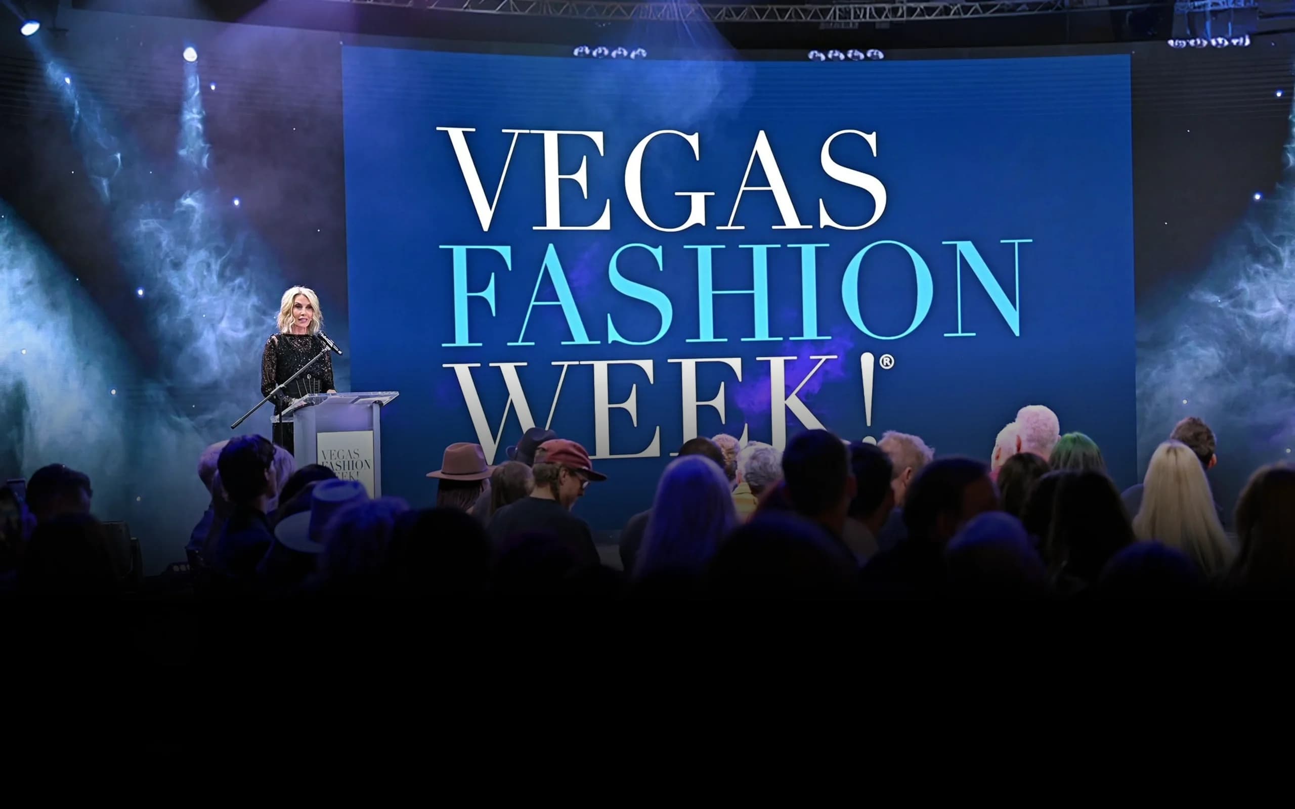 A speaker addressing the audience at Vegas Fashion Week®, with the event's name shown on a stage screen behind them